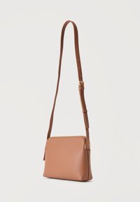 Μικρή καφέ δερμάτινη τσάντα χιαστί με ρυθμιζόμενο λουράκι και χρυστό φερμουάρ, απεικονίζεται σε απλό φωτεινό φόντο.