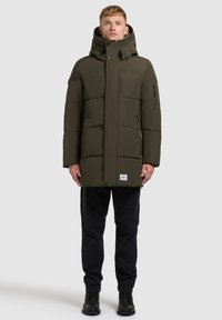 Donkergroene, gewatteerde parka met een hoge kraag en capuchon. Voorzien van ritszakken aan de zijkant en een logopatch aan de zoom. Gecombineerd met zwarte broek en laarzen.