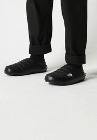 Black quilted slip-on shoes with a textured surface and a rubber sole, featuring a white logo on the side, worn with black pants.