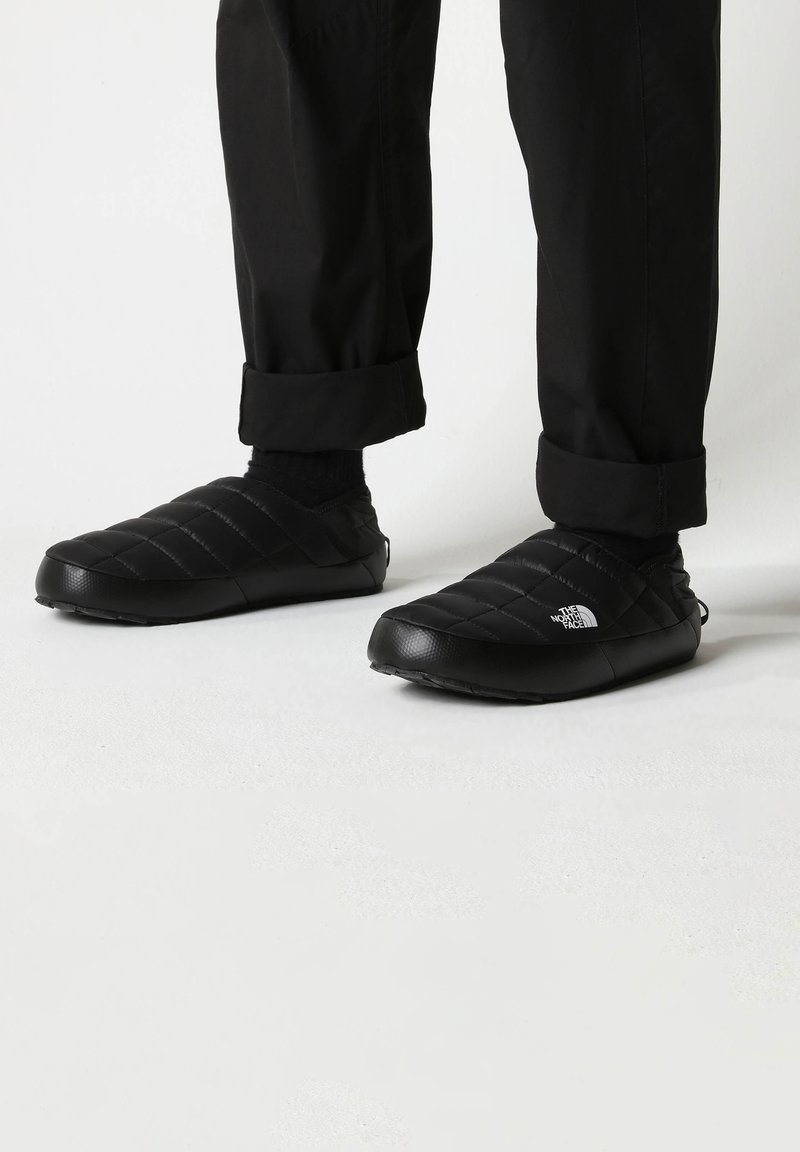 Black quilted slip-on shoes with a textured surface and a rubber sole, featuring a white logo on the side, worn with black pants.