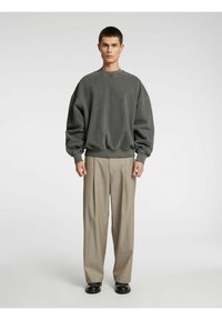 Pelēks pārlielizmēra džemperis ar ribveida aprocēm, kombinēts ar bēšām, plaša piegriezuma biksēm. Modelis stāv neitrāla fonā.