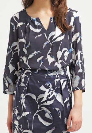 Blouse marine avec motif floral blanc ; présente un col en V, des manches 3/4 et une coupe ample. Fabriquée en tissu léger avec un léger éclat.