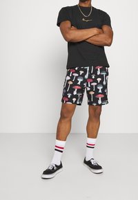 T-shirt nera con scritta dorata, abbinata a pantaloni corti neri con un motivo di funghi colorati, calze bianche con strisce rosse e nere, e sneaker nere.