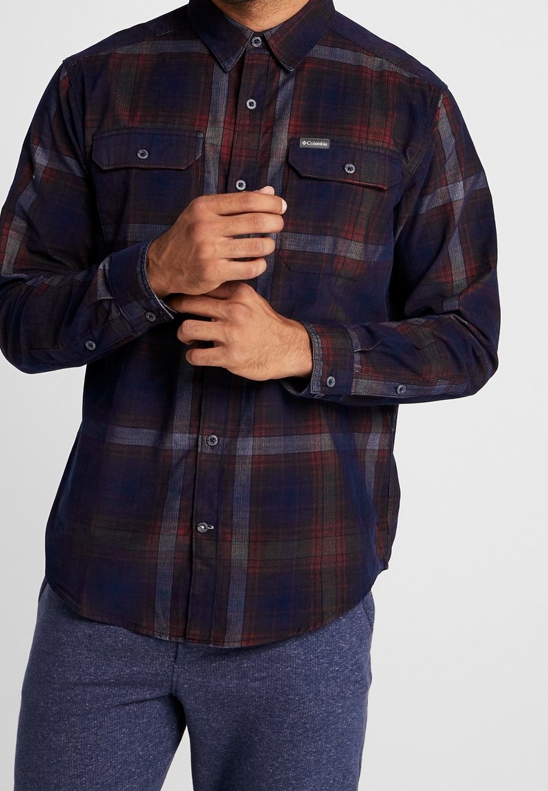 Homme portant une chemise à carreaux foncée boutonnée avec des poches poitrine et un pantalon bleu, ajustant une manchette avec ses deux mains.