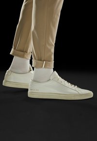 Białe skórzane sneakersy z zaokrąglonymi noskami, teksturowanymi podeszwami i subtelnym tłoczeniem po zewnętrznej stronie, połączone z podwiniętymi beżowymi spodniami i białymi skarpetkami.