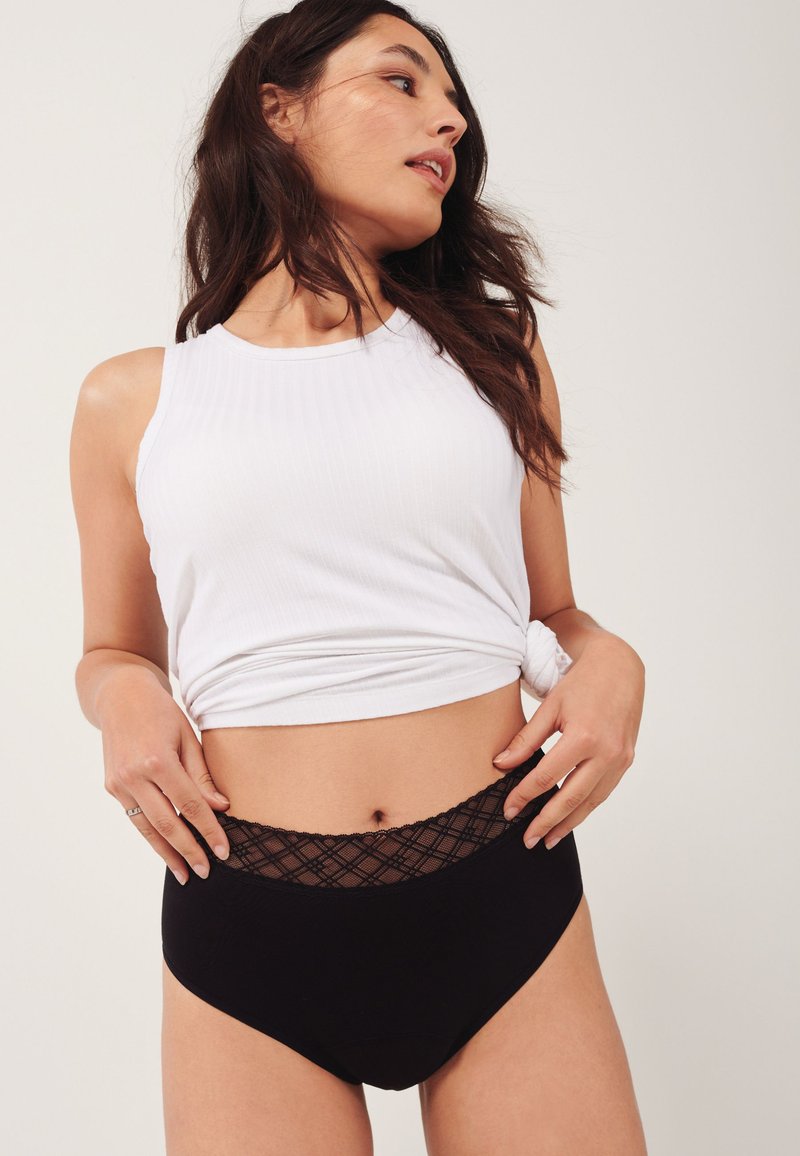 Femme portant un haut blanc sans manches noué à la taille et un short taille haute noir avec une ceinture en dentelle, posant devant un fond uni.