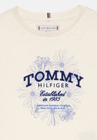 Κοντινό πλάνο μπλούζας Tommy Hilfiger σε κρέμα απόχρωση με μπλε λουλουδάτο γραφικό και κείμενο "Ιδρύθηκε το 1985" στο κέντρο μπροστά.