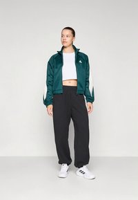 Casaco de cetim verde com fecho e detalhes brancos, combinado com calças pretas de corte largo e sapatilhas brancas. Apresenta um logótipo da adidas no peito.