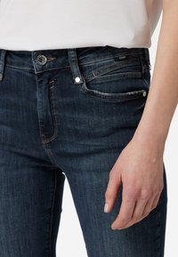 Jeans de mezclilla azul oscuro con un ajuste slim. Las características incluyen un cierre frontal con botón, detalles metálicos y costuras visibles.