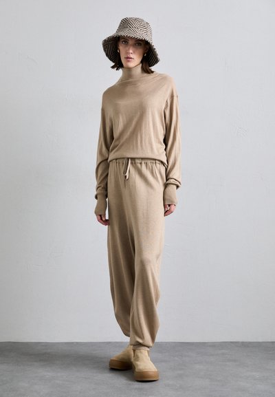 Beige knitted sweater and jogger set with a high neck and elastic waistband, paired with a patterned bucket hat and chunky-soled boots.