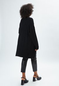 Cardigan oversize in maglia nera a trecce con spalle scese, abbinato a pantaloni cropped in pelle nera e scarpe con tacco.