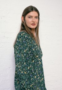 Vestido com estampa floral em verde, apresentando pequenas flores amarelas e roxas, mangas longas e design em V. Tecido suave com uma forma fluida.