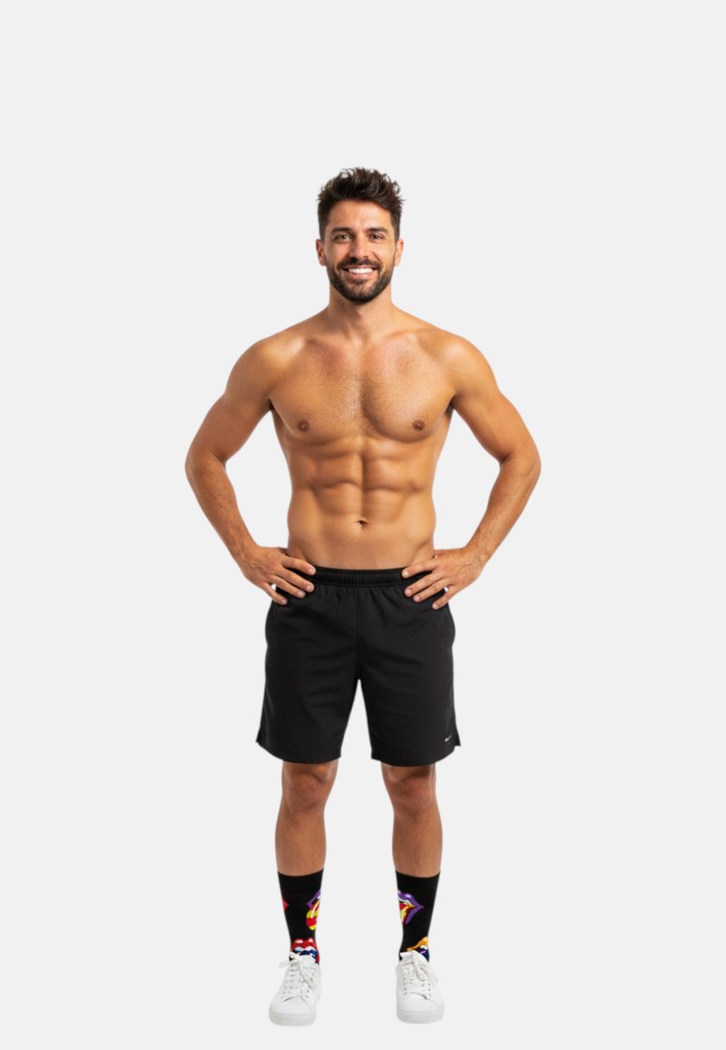 En mand med en muskuløs bygning står med hænderne på hoften iført sorte atletiske shorts, farverige mønstrede sokker og hvide sneakers.