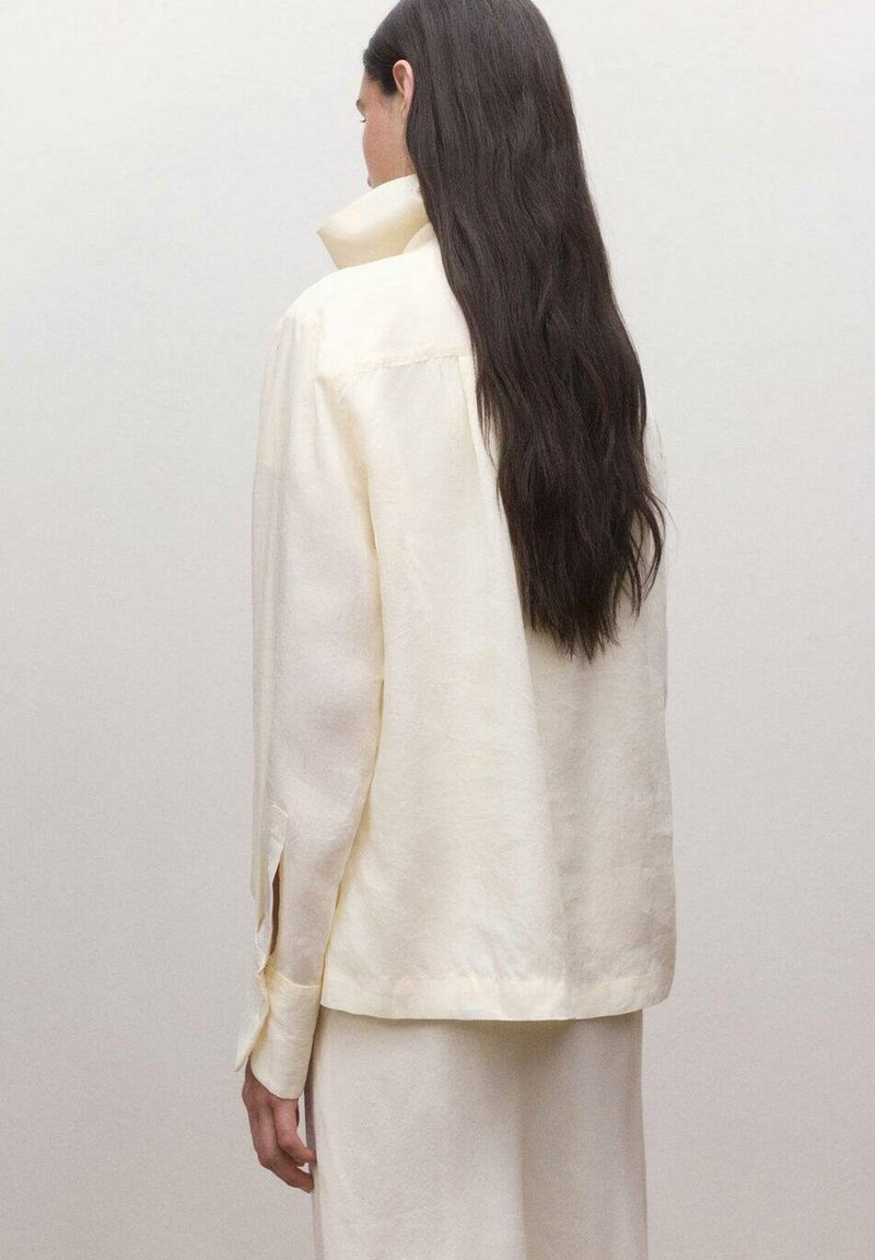 Personne aux cheveux longs et foncés portant une veste ample de couleur crème avec les manches boutonnées aux poignets, debout devant un arrière-plan clair uni.