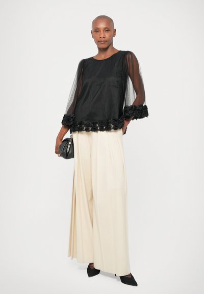 Black sheer top with floral lace trim, paired with cream wide-leg trousers and black pointed shoes. Black clutch bag completes the look.