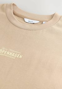 T-shirt en coton beige avec un col rond. Présente un texte en relief "COPENHAGUE" en blanc, sur un tissu texturé. Coupe décontractée.