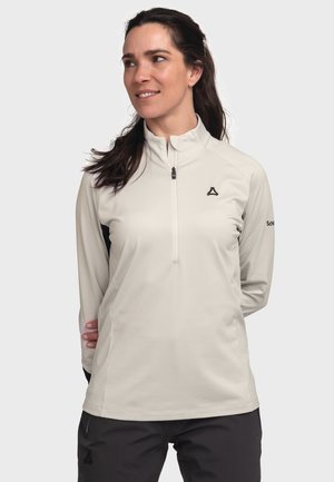 Woman with dark hair wearing a light gray long-sleeve zip-up shirt and dark pants standing against a plain background.