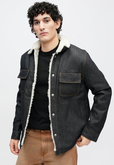 Tmavě modrá džínová bunda s bílým umělým kožešinovým límcem. Má zapínání na knoflíky a dvě náprsní kapsy. Hnědé švy zdůrazňují švy.