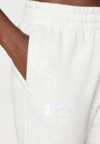 Bijele flis trenirke s elastičnim pojasom, bočnim džepovima i suptilnom teksturom. Sadrže bijeli vezeni Under Armour logo.
