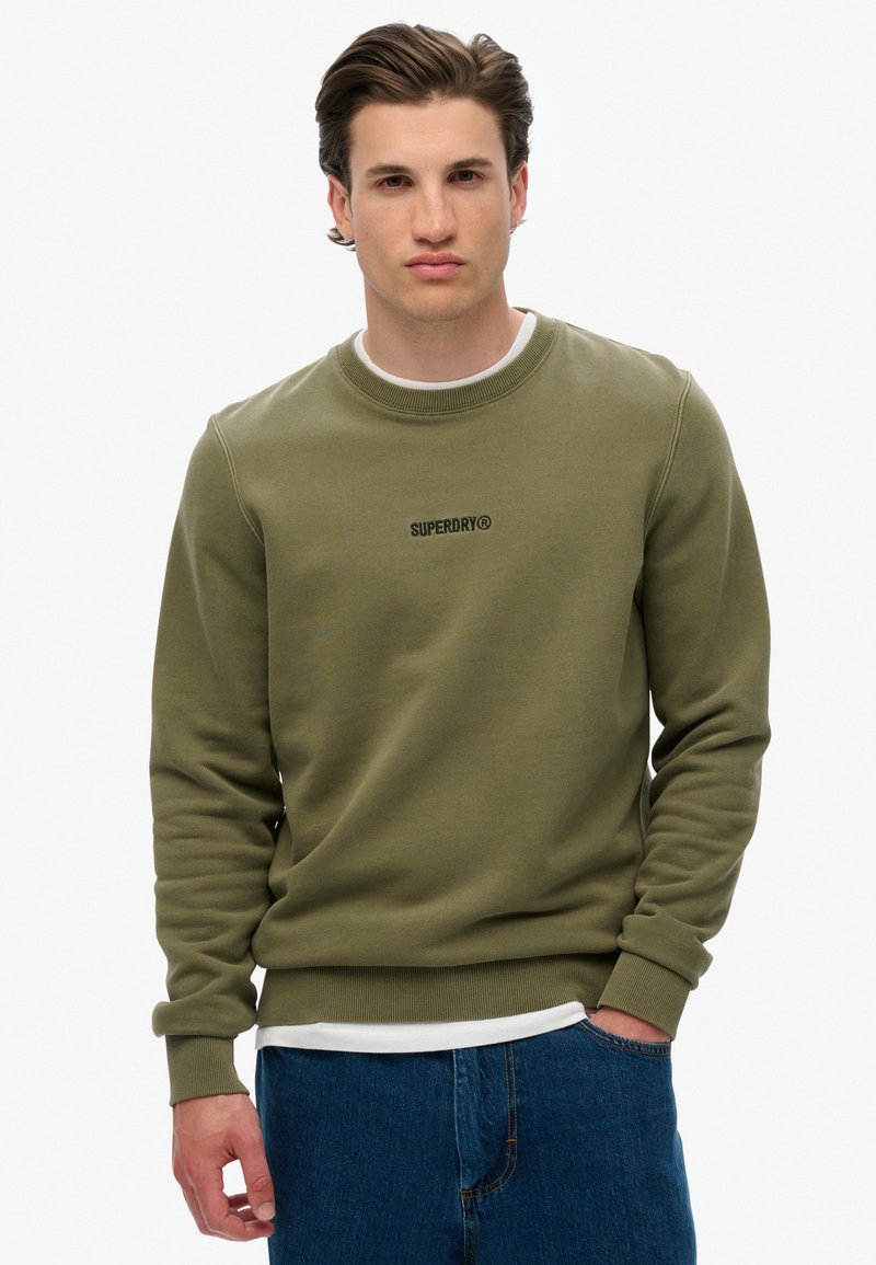 Sudadera verde oliva con textura acanalada, cuello redondo y un pequeño logo negro en el pecho. Se lleva sobre una camisa blanca y combinada con jeans azules.