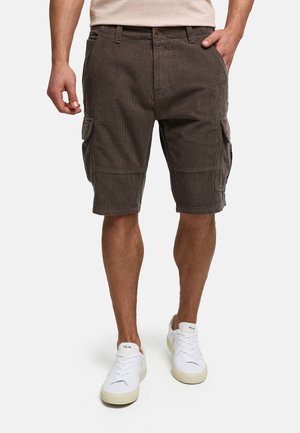 Mand iført mørkebrune cargo-knælange shorts og hvide Veja-sneakers, stående med den ene hånd i lommen mod en ensfarvet baggrund.