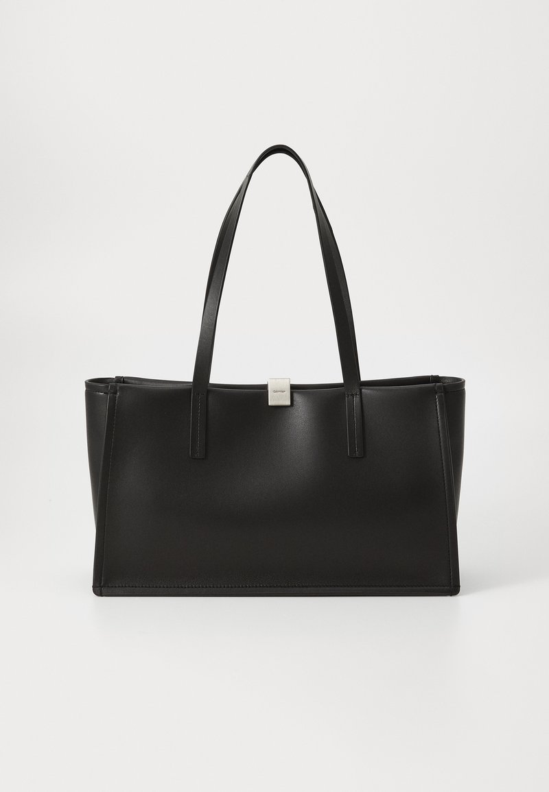 Črna sintetična torba s sleek dizajnom, ravnim dnom in dvema dolgima ročajema. Na sprednji strani se nahaja majhna srebrna oznaka.