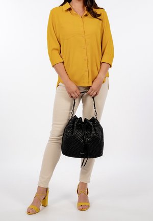 Frau trägt eine senfgelbe Bluse und beige Hose, hält eine schwarze gewebte Bucket-Bag und trägt passende senfgelbe Sandalen mit Absatz.