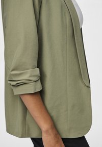 Olivgrön kavaj med uppvikta ärmar, med en slät yta, öppen front och minimalistisk design utan knäppningar eller mönster.