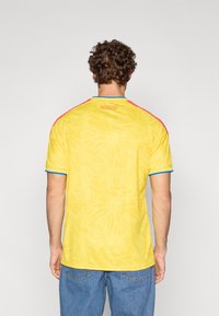 Camisola desportiva amarela com textura padrão, gola azul e detalhes, ombros vermelhos e "COLOMBIA" impresso na parte de trás do pescoço.