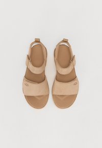 Beige mokkasandaalit, joissa on säädettävät Velcro-hihnat, pehmustettu pohja ja kumipohja. UGG-logo painettuna sivulle.