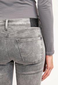 Jeans de mezclilla gris de corte ajustado, con bolsillos traseros, costuras en contraste y un parche de cuero negro en la cintura.