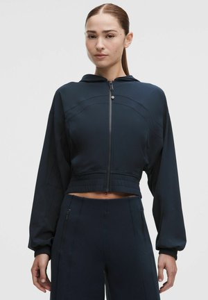 Ung kvinde med sat hår tilbage iført en mørk marineblå cropped zip-up hoodie og matchende højtaljede bukser med lynlåslommer.