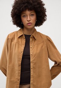 Camicia di corduroy marrone con chiusura a bottoni, colletto a punta e maniche a sbuffo, indossata sopra un top fitted marrone scuro.