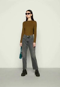 Καφέ ριγέ μακρυμάνικο τοπ, γκρι ψηλόμεσα τζιν, μαύρες χοντρές μπότες και μια τσάντα σε κυπαρισσί χρώμα. Το μοντέλο στέκεται μπροστά σε ένα απλό φόντο.