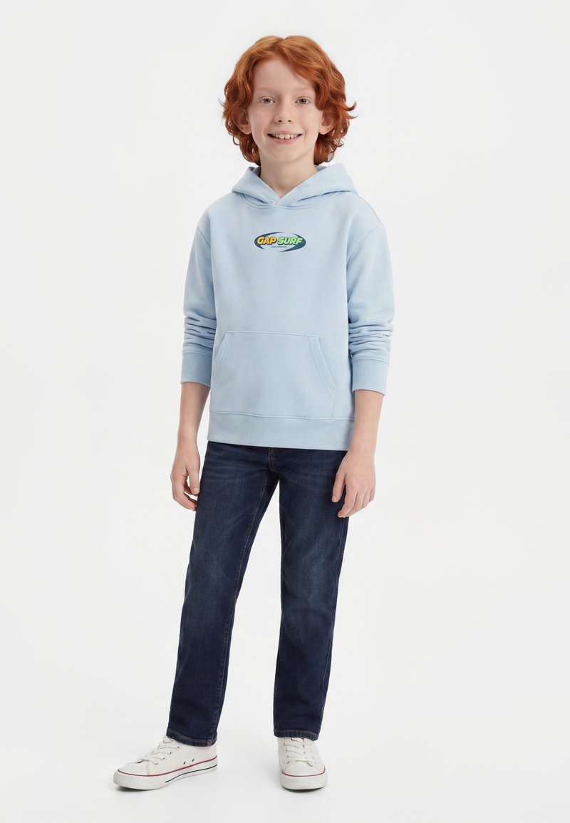 Enfant souriant aux cheveux roux, portant un sweat à capuche bleu clair GAP SURF, un jean foncé et des baskets blanches, debout devant un fond blanc uni.