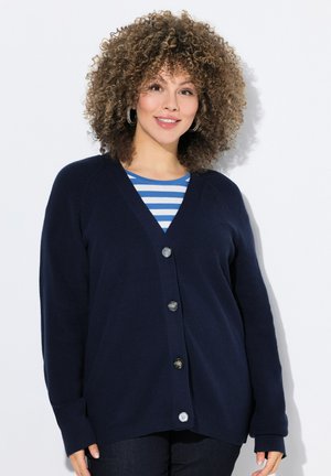 Cardigan bleu marine avec un col en V, fermeture à cinq boutons, texture côtelée et manches longues. Porté sur un haut rayé bleu et blanc.