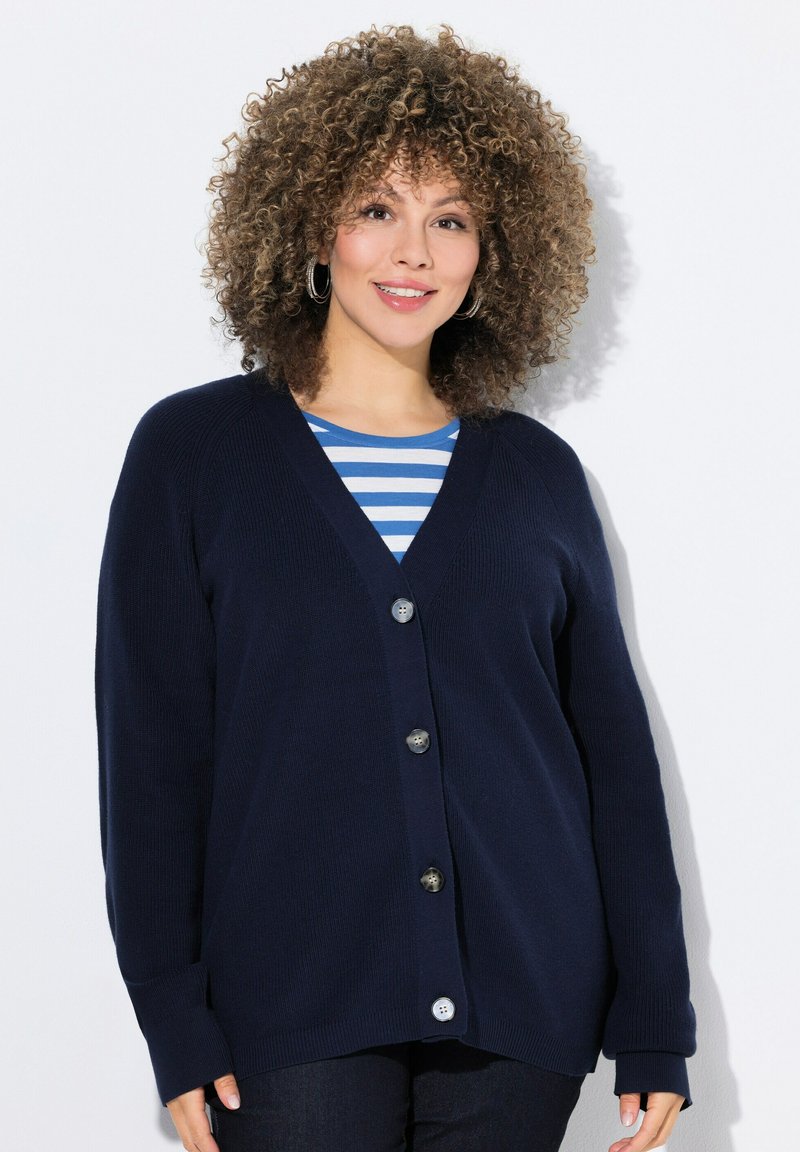 Cardigan bleu marine avec un col en V, fermeture à cinq boutons, texture côtelée et manches longues. Porté sur un haut rayé bleu et blanc.