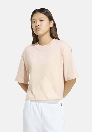 Joven con el pelo largo y oscuro que lleva una camisa ligera de manga corta de color rosa claro y pantalones blancos sobre un fondo liso.