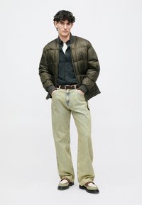 Noor mees, kes kannab oliivrohelist sulejacketit, musta särk, heledat bežist laia jalaga teksapükse, pruuni vööd ja beezide ja pruunide kingadega, seisab käed taskutes.