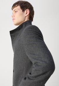Grau kariierter Mantel mit hohem Kragen, der eine taillierte Silhouette, strukturierten Stoff und einen vorderen Knopfverschluss aufweist.