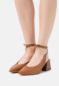 Talons en cuir tressé marron avec un bout pointu, une ouverture à l'arrière et des lacets à la cheville. Des talons en bois épais et une texture lisse complètent le design.