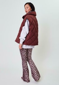 Pletená hnedá vesta s vysokým golierom, nosená cez šedú mikinu, v kombinácii s rozšírenými nohavicami s leopardím vzorom. Viditeľné sú hladké štruktúry látok.