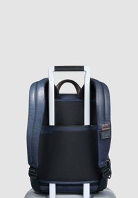 Mochila azul marino con un exterior liso y un panel negro texturizado, equipada con correas acolchadas y un asa superior. Diseñada para colocarse sobre los tiradores del equipaje.