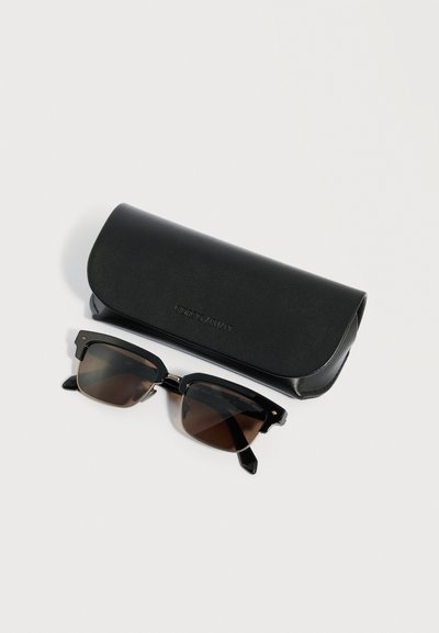 Črna pravokotna sončna očala z temnimi lečami ob črni trdi torbici z reliefnim napisom, postavljeni na enostavno belo površino.