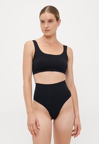 Woman wearing black high-waist underwear and a black crop tank top with lace trim, standing against a plain white background.