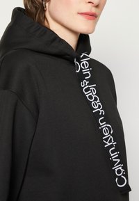 Felpa nera con cappuccio e coulisse, caratterizzata dal logo bianco "Calvin Klein". Texture morbida e liscia, con vestibilità rilassata e design minimalista.