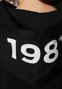 Svart huvtröja med stort vitt "1981" tryckt på baksidan, bärs av en person med brunt hår synligt vid nacken.