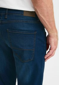 Jeans oscuros de mezclilla, con un bolsillo trasero que presenta costuras amarillas en contraste y una etiqueta marrón en la cinturilla.