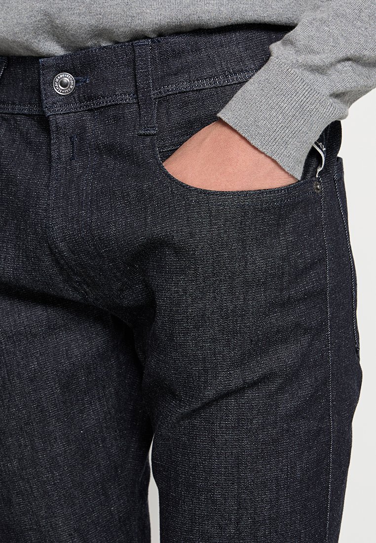 Dunkelblaue Jeans im Slim-Fit-Schnitt mit Fronttaschen, kontrastierenden Nähten und einem Reißverschluss mit einem Metallknopfdetail.