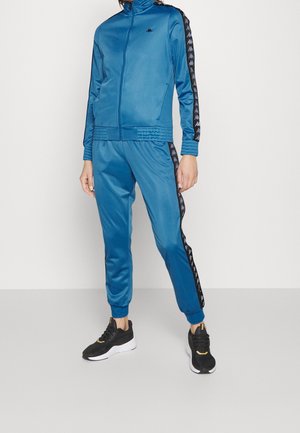 Osoba w turkusowym dresie z czarnymi paskami z logo na rękawach i bokach, w czarno-białych trampkach, stojąca na białym tle.